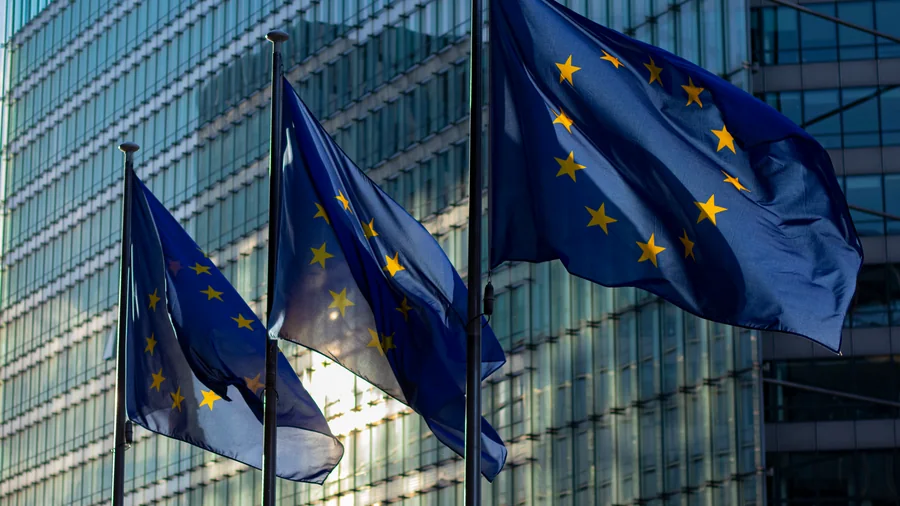 Drie Europese vlaggen wapperen aan masten voor een glazen kantoorgebouw