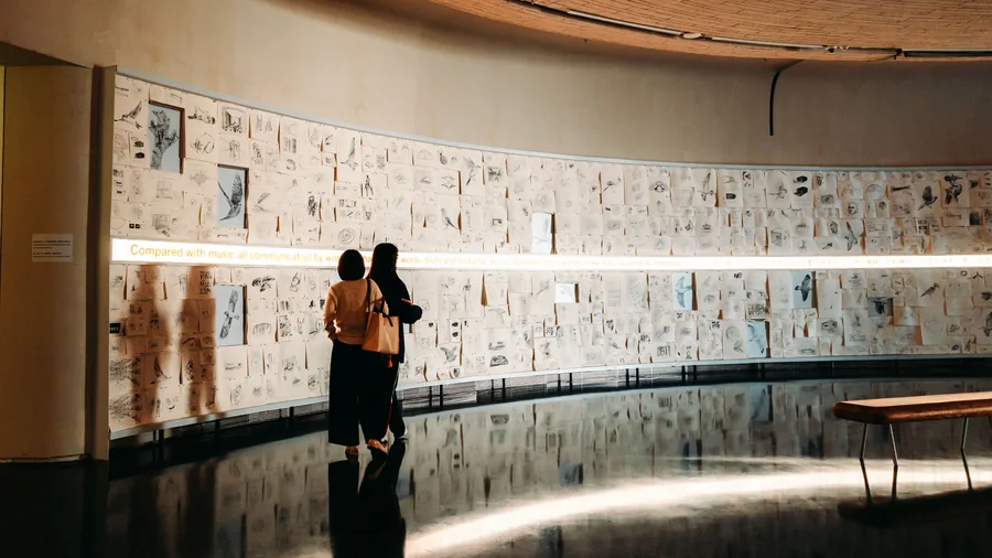 Twee bezoekers lopen langs een gebogen wand vol tekeningen in een museumzaal