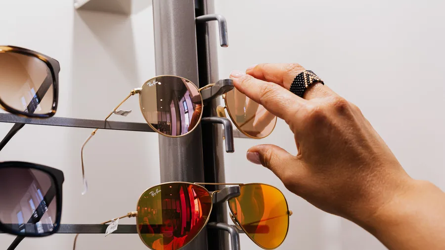 Hand pakt een Ray-Ban-zonnebril met spiegelende glazen van een rek in een brillenwinkel