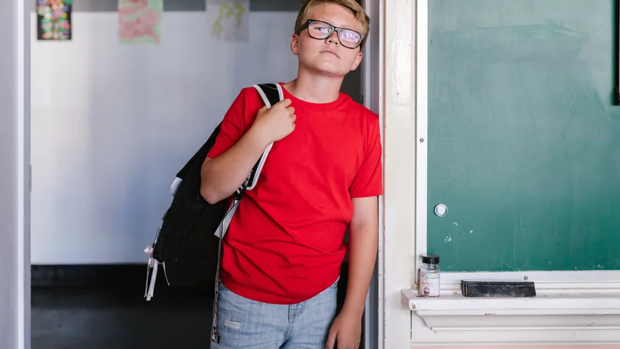 Een jongen met een bril en een rugzak staat bij het schoolbord in een klaslokaal