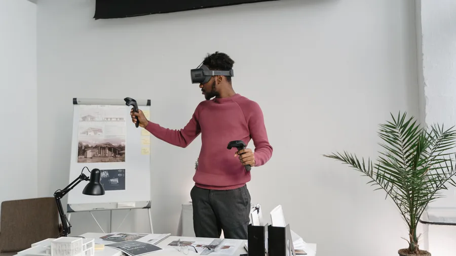 Man met een VR-bril en twee controllers staat in een lichte kamer naast een tafel met papieren