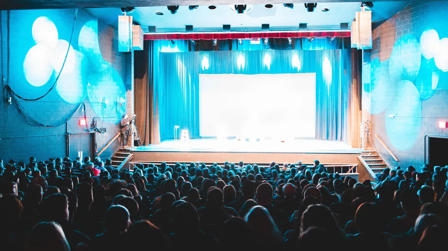 Publiek zit in een volle theaterzaal en kijkt naar een verlicht podium met een blauw doek