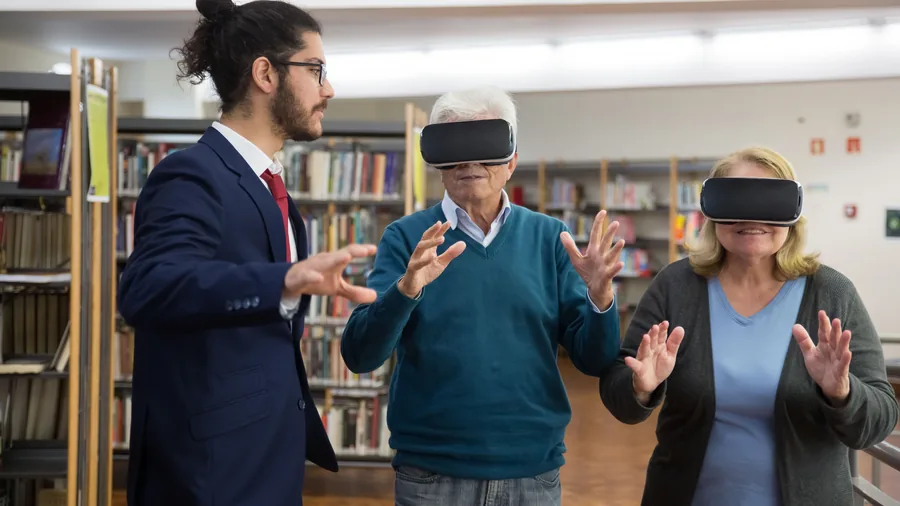 Een oudere man en vrouw met een VR-bril op staan in een bibliotheek terwijl een jonge man naast hen uitleg geeft