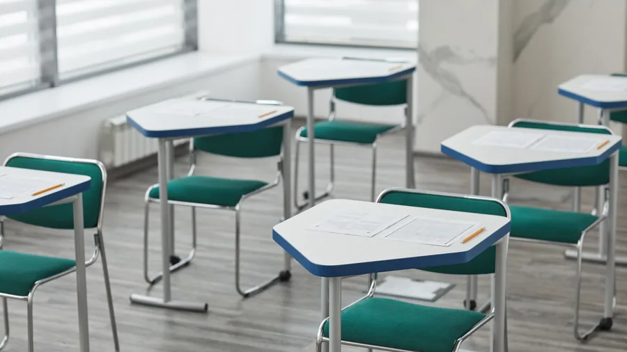 Lege witte schooltafels met examenpapieren en groene stoelen in een lichte klas met lamellen voor de ramen