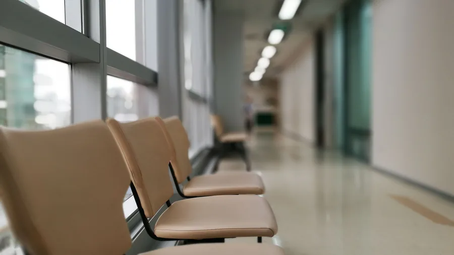 Een rij lege beige wachtkamerstoelen in een lichte ziekenhuisgang met ramen